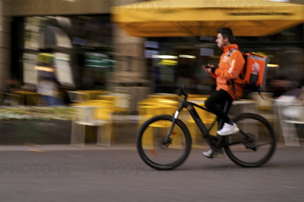 Lieferando, logo, delivery service, courier service, bicycle courier, courier driver rides with thermal backpack, e-bike, mobile phone, smartphone, movement effect, speed, blurred, Stuttgart, Baden-Württemberg, Germany