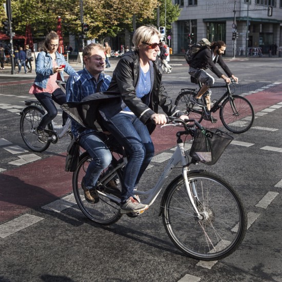 DEU Germany Germany Berlin cyclist transports a man on the luggage rack while another woman watches her smartphone. Everyone is driving on the road 'Unter den Linden' towards Brandenburg Gate