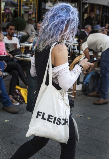 DEU Germany Germany Berlin Woman with blue-dyed hair wears a cloth bag with the inscription Flesh while she is strolling into her smartphone