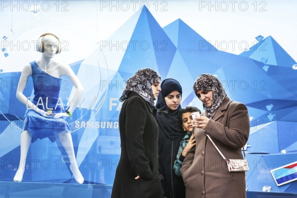DEU Germany Germany Berlin Women wearing headscarves look at an Apple iPhone. You are standing in front of a KaDeWe shop window, in which Korean electronics manufacturer Samsung is promoting its Galaxy smartphone