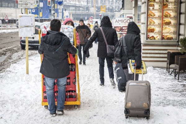 DEU Germany Germany Berlin Travelers with wheeled suitcases pass by beverage purveyors on a snow-covered sidewalk near Zoo Station