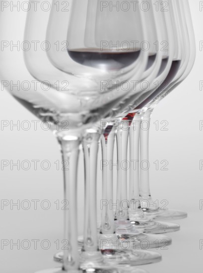Some wine glasses standing consecutive in a row in light grey back, one partly filled with red wine