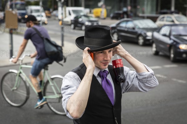 DEU Germany Germany Berlin A man protects his hat from a gust of wind while talking on the phone and holding a coke bottle