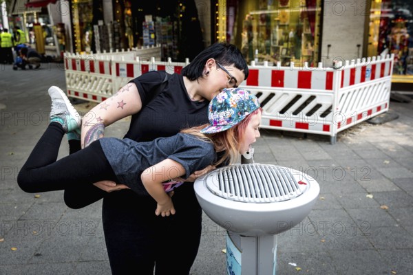 DEU Germany Germany Berlin A woman holds a girl over a stream of water gushing from a public water dispenser on Kurfürstendamm. Berlin's waterworks offer free drinking water at numerous locations in the city