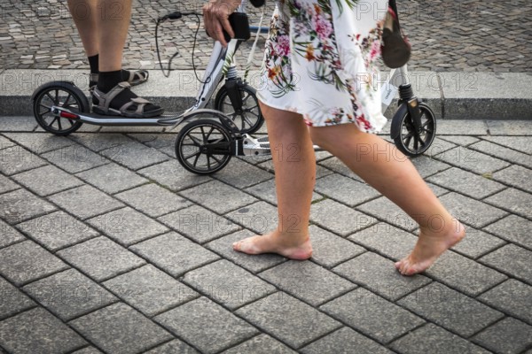 DEU Germany Germany Berlin A woman passes a family on pedal scooters barefoot on Pariser Platz in front of the Brandenburg Tor