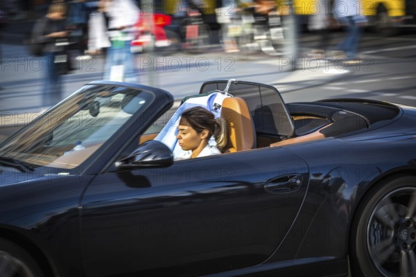 DEU Germany Germany Berlin A woman is dazzled by the sun as she drives her convertible across Potsdamer Platz. She has a change of clothes hanging on the windscreen