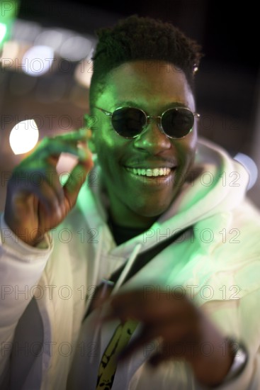 Greenlit African man wearing sunglasses at night as shade of anonymity, approved immigration, integration, laughing, happy, giggle, funny, joyful, optimistic, anticipation, expecting, expectation, optimism, proactive, can do attitude, outgoing, charming, big smile, open-minded, optimist, immigrant, foreigner, approval, green, environmentalism, environmental attitude, sustainability, green as color for sustainable environment, candid happiness, jolly, excited, excitement, high spirited, elated, motivated, motivation, good mood, movement, cheerful, cheerfulness