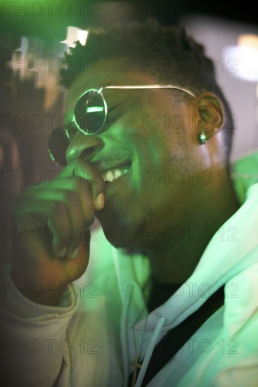 Greenlit African man wearing sunglasses at night as shade of anonymity, laughing, happy, giggle, funny, joyful, optimistic, optimist, approved immigration, integration, immigrant, foreigner, approval, green, environmentalism, environmental attitude, sustainability, green as color for sustainable environment, amusement, amused