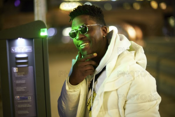 Thoughtful greenlit African man at parking ticket machine pausing as symbol for approved immigration and ability to pause and recharge, wearing sunglasses as shade of anonymity, scepticism, thinking, pondering, thoughts, scpetic, sceptical, hesitation, hesisting, hand on chin, integration, smile, smiling, amused, good mood, optimist, optimistic, optimism
