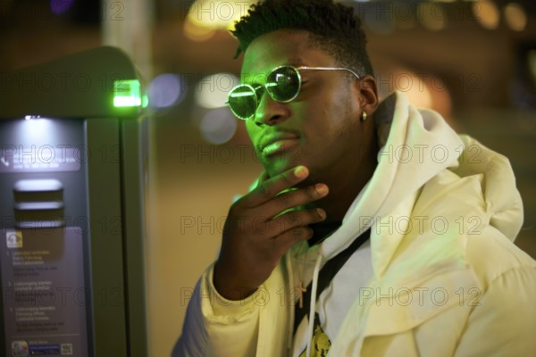 Thoughtful greenlit African man at parking ticket machine pausing as symbol for approved immigration and ability to pause and recharge, approval, immigrant, wearing sunglasses as shade of anonymity, scepticism, thinking, pondering, thoughts, scpetic, sceptical, hesitation, hesisting, hand on chin, integration, green as color for sustainability and environmentalism, sustainable mobility transition, serious attitude