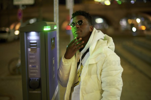 Thoughtful greenlit African man at parking ticket machine pausing as symbol for approved immigration and ability to pause and recharge, immigrant, wearing sunglasses as shade of anonymity, scepticism, thinking, pondering, thoughts, scpetic, sceptical, hesitation, hesisting, hand on chin, approval, integration, green as color for sustainability and environmentalism, sustainable mobility transition