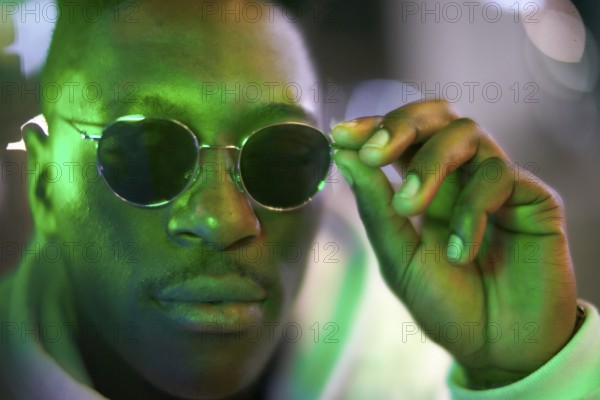 Greenlit African man wearing sunglasses at night as shade of anonymity, coolness, approved immigration, integration, immigrant, foreigner, approval, green, environmentalism, environmental attitude, sustainability, poised, serious, calm, close-up, green as color for sustainable environment and optimism and being greenlit approved
