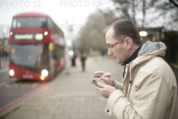 Man typing on smartphone next to double-deck bus in London, red bus, checking timetable on mobile phone touching touchscreen with pointer finger using app navigating his city commute travels in London, Great Britain, bus stop, bus is stopping, waiting, daily life, Northern European ethnicity, street traffic, app, internet, sightseeing, digital, navigation, searching, connection, public transportation, route, public transport, waiting