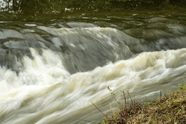 Borken-Hoxfeld, NRW, Germany, Water cascades smoothly over rocks, creating gentle waves along the riverbank. Lush green vegetation lines the edge, adding to the peaceful atmosphere on a sunny afternoon