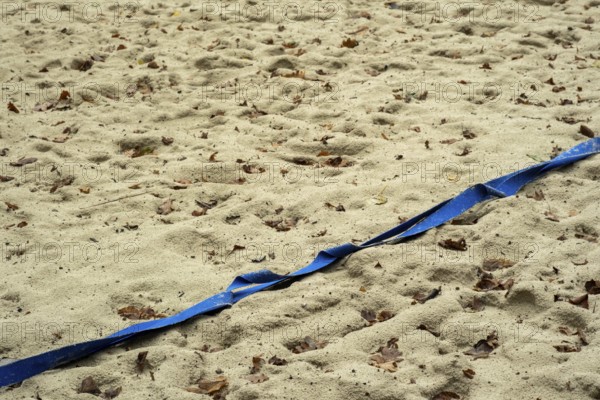 Borken-Hoxfeld, NRW, Germany, A stretch of sandy beach shows blue tape running across, marking a section. The ground is covered in fallen leaves, suggesting a recent autumn day. Footprints indicate activity