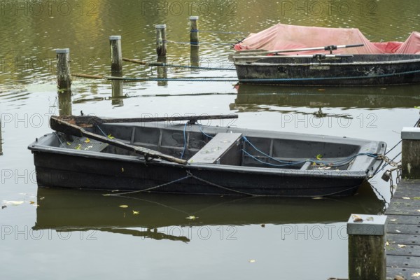 Borken-Hoxfeld, NRW, Germany, A weathered black boat is moored at the dock, surrounded by tranquil water. Nearby, another small boat is visible. Fallen leaves float on the surface under an autumn sky