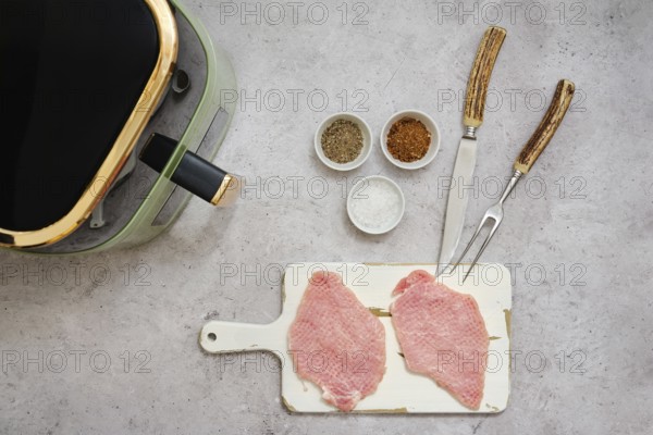 Two raw pork sirloin cutlets are placed on a cutting board next to small bowls filled with spices. An air fryer sits nearby with utensils ready for cooking. The scene is set for a delicious meal
