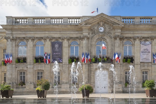 The classicist façade of Beauvais Town Hall (Hotel de Ville) dominates Place Jeanne Hachette. Floor fountains can be seen in action in the foreground. The building is decorated with numerous tricolor flags (drapeaux francais), under a cloudy blue sky in Beauvais, Oise department (60), Hauts-de-France region, France