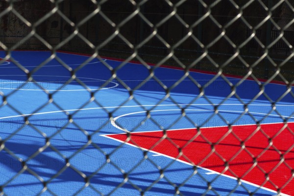 Brightly colored basketball court features blue and red sections, seen through a chain-link fence. Sunlight casts shadows on the court, indicating a sunny day in the city
