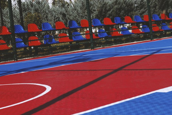 A brightly colored basketball court features bold red and blue sections under the clear sky. Nearby, empty chairs line the sidelines, ready for spectators on a sunny day