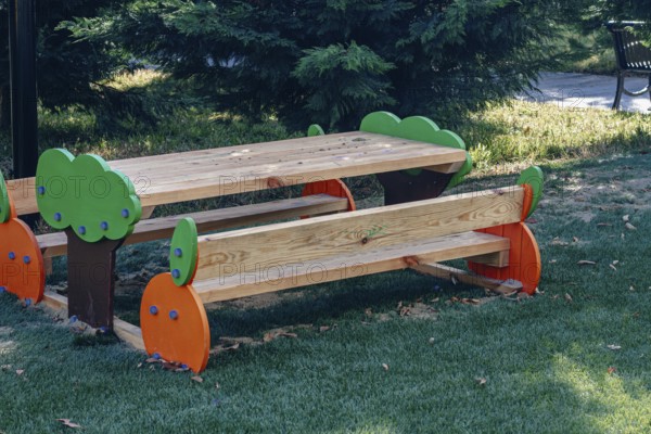 A vibrant wooden picnic table with colorful tree designs stands in a green park, surrounded by grass and trees under clear blue skies on a sunny day
