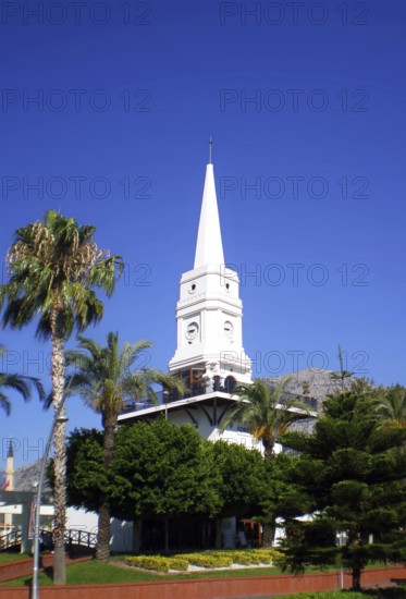 A tall white clock tower surrounded by palm trees, one of the main architectural landmarks for tourists and travelers of the resort town of Kemer (Antalya Province) and the entire eastern Mediterranean Turkish Riviera. Kemer, Turkey