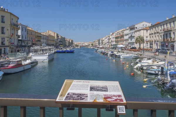 The Canal de Sete, formerly Canal Royal, stretches between the historic house facades, with an information board on the history of the canal in the foreground and numerous moored boats on the banks, seen from the Pont de la Savonnerie under a blue sky in Sete, Herault department, Occitanie region, France