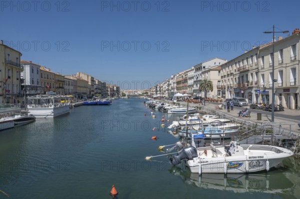 The wide Canal de Sete (formerly Canal Royal) is surrounded by historic house facades, with numerous moored motor boats on Quai Leopold Suquet, seen from the Pont de la Savonnerie under a blue sky in Sete, Herault department, Occitanie region, France