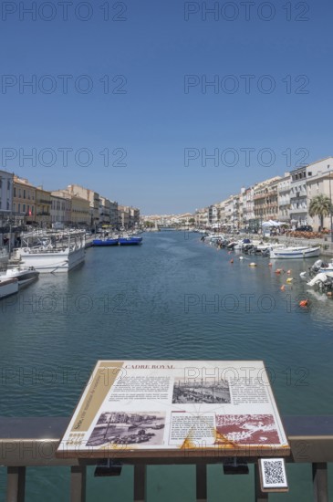 The Canal de Sete, formerly Canal Royal, stretches between the historic house facades, with an information board on the history of the canal in the foreground and numerous moored boats on the banks, seen from the Pont de la Savonnerie under a blue sky in Sete, Herault department, Occitanie region, France