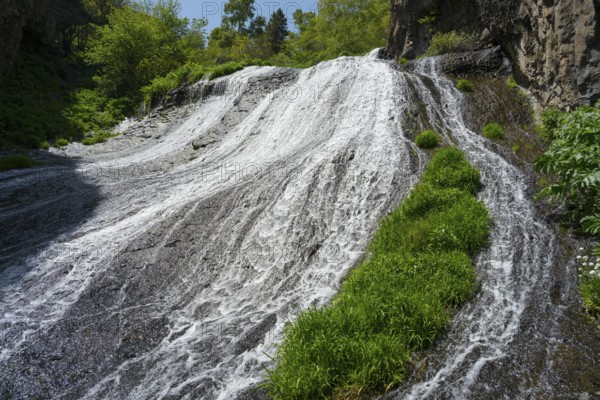 A wide waterfall flows steeply down, flanked by green plants and rocks in nature, Jermuk Waterfall, Vayots Dzor Province, Armenia