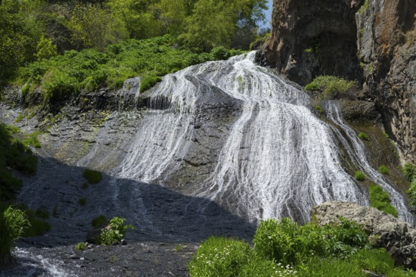 A clear waterfall flows over rocks surrounded by lush green plants in sunny nature, Jermuk Waterfall, Vayots Dzor Province, Vayots Dzor, Armenia