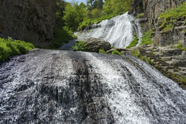 A powerful waterfall pours over steep rocks surrounded by green vegetation in the sun, Jermuk Waterfall, Jermuk, Vayots Dzor Province, Armenia