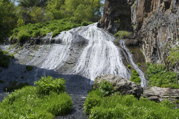 A bright waterfall snakes through green plants and rocks in sunny nature, Jermuk Waterfall, Vayots Dzor Province, Armenia