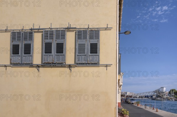 Yellow house façade with three gray wooden shutters to move and a view across the Canal de Sete (formerly Canal Royal) to the distinctive steel turnstile Pont Marechal Foch and the port area in La Pointe Courte in Sete, Herault department, Occitanie region, France