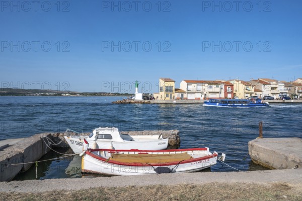 Two moored fishing boats in the foreground with a view across the Canal de Sete, formerly Canal Royal, of the colorful houses and the white-green lighthouse of La Pointe Courte, while a blue sightseeing boat passes by, under a blue sky in Sete, Herault department, Occitanie region, France