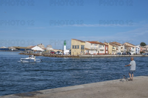 An angler looks from the banks of the Canal de Sete, formerly Canal Royal, across to the green-white lighthouse of La Pointe Courte, while a man steers his motorboat out onto the Etang de Thau, Sete, Herault department, Occitanie region, France