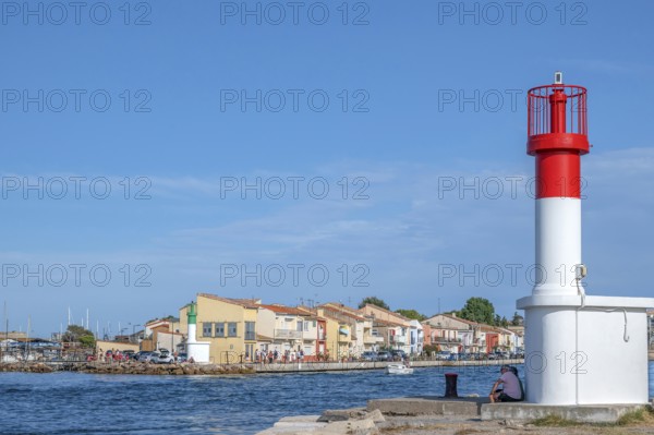 Men sitting at the red and white lighthouse at the port entrance of the Canal de Sete, formerly Canal Royal, with the typical, colorful fishermen's houses of the La Pointe Courte quarter under a blue sky in Sete, Herault department, Occitanie region, France