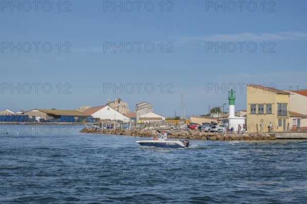 A motorboat with people on board steering it sails in front of the white-green little lighthouse and a yellow fisherman's house at the port entrance of the Canal de Sete, formerly Canal Royal, at La Pointe Courte with industrial buildings in the background under a blue sky in Sete, Herault department, Occitanie region, France