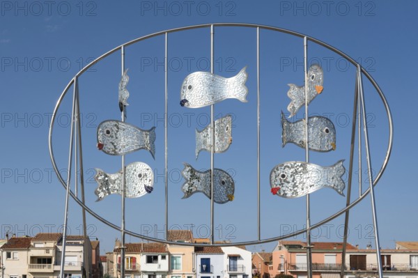 The modern metal sculpture Les Daurades Miroirs by Dominique Dore on the Plage de la Lagette shows silver mirror fish in an oval frame, with the colorful houses of the La Pointe Courte district in Sete, Herault department, Occitanie region, France