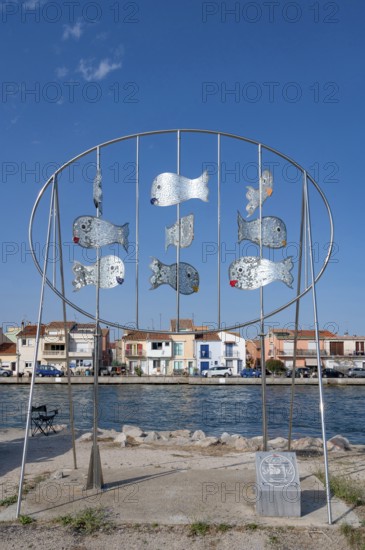The modern metal sculpture Les Daurades Miroirs by Dominique Dore on the Plage de la Lagette shows silver mirror fish in an oval frame, with the colorful houses of the La Pointe Courte district in Sete, Herault department, Occitanie region, France