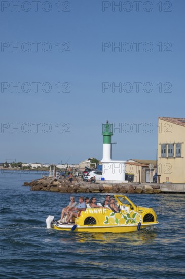 Sightseeing on the water: a yellow water car with tourists and lemon decorations rides on the Canal de Sete, formerly Canal Royal, in front of a white-green lighthouse and fishermen's houses in La Pointe Courte under a bright blue sky in Sete, Herault department, Occitanie region, France