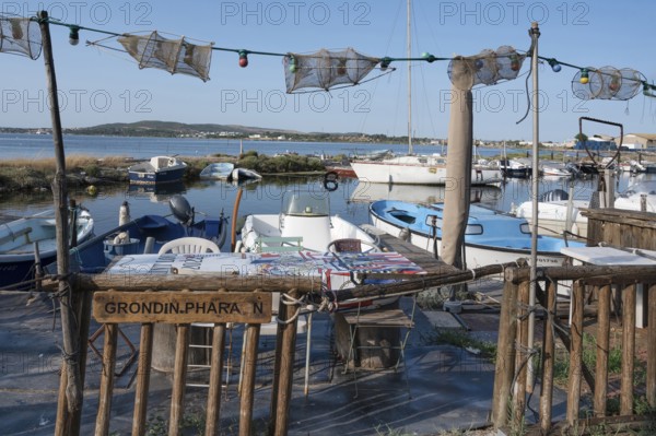 A fisherman's sitting area for an aperitif with tables and chairs as well as fishing nets as decoration, framed by fishing boats in the small port of La Pointe Courte with a view of the Etang de Thau under a blue sky in Sete, Herault department, Occitanie region, France