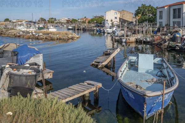 A blue fishing boat is moored on dilapidated wooden walkways in a small harbor basin of La Pointe Courte, surrounded by fishing equipment and baskets with houses and other boats in the background under a blue sky in Sete, Herault department, Occitanie region, France
