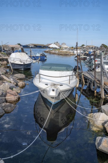 A white moored motor boat with reflection in the calm water of a small harbor of the traditional fishing port of La Pointe Courte between rocks and wooden walkways, with other fishing boats and industrial buildings under a blue sky in Sete, Herault department, Occitanie region, France