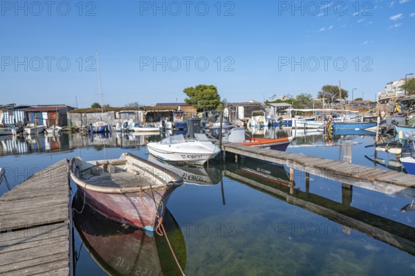 A red-faded fishing boat is moored on a wooden pier in the port of La Pointe Courte, whose reflection is perfectly reflected in the calm water, surrounded by other boats and the colorful fishing huts in the background under a clear blue sky in Sete, Herault department, Occitanie region, France