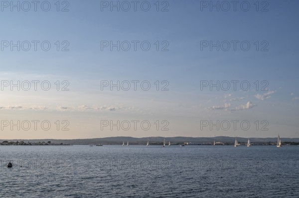 Wide-angle landscape view of Etang de Thau with several sailboats on the water sailing towards the skyline of the Balaruc coast, seen from the banks of the Le Barrou district under blue summer skies in Sete, Herault department, Occitanie region, France