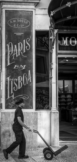 Black and white photograph, street art, man with a sack truck at the entrance to a restaurant, Lisbon, Portugal