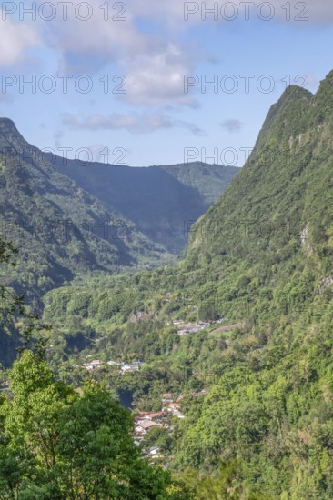 View from Department 52 of the massive, steep Remparts rock faces of Cirque du Salazie and a hamlet at the bottom of the valley on a sunny valley with some clouds in the blue sky, Cirque de Salazie, Tropical Highlands, La Reunion, France