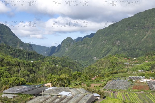 Tropical valley in Cirque de Salazie with intense green Rempart cliffs and slopes surrounded by low-hanging clouds under a blue sky, while agriculture, wax houses and houses shape the low-lying valley floor in the foreground, Cirque de Salazie, Tropical Highlands, La Reunion, France