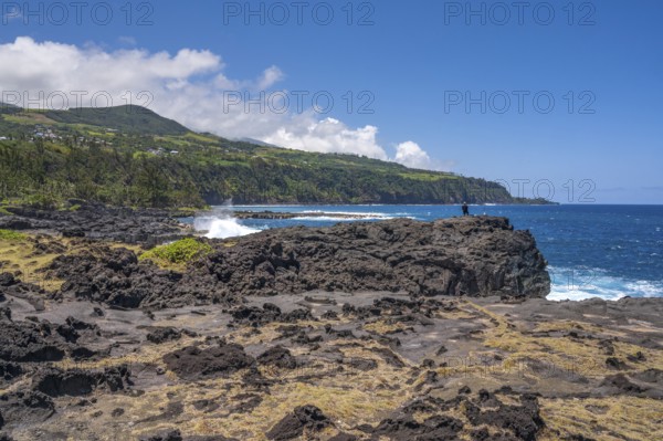 An angler stands on a massive rock of the black lava coast and looks out at the roaring Indian Ocean as the surf white washes around the volcanic rock and in the background the steep, green cliffs near the Marine de Vincendo rise into the sky, Marine de Vincendo, South Coast, La Reunion, France